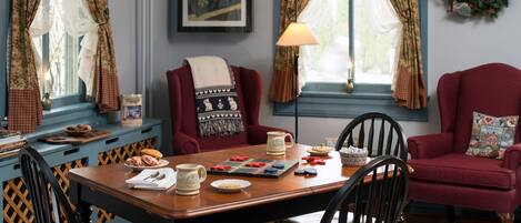 Un coin repas chaleureux avec une table en bois, des chaises et un fauteuil rouge. Une lampe et un tableau encadré décorent les murs.