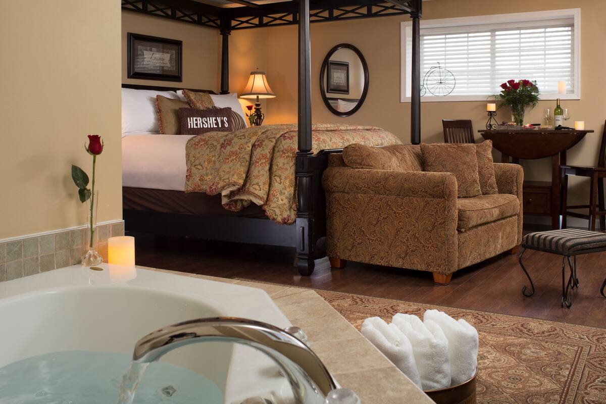 A bedroom with a large bed, a sitting area with a patterned sofa, a small table with a vase, and a window with blinds.