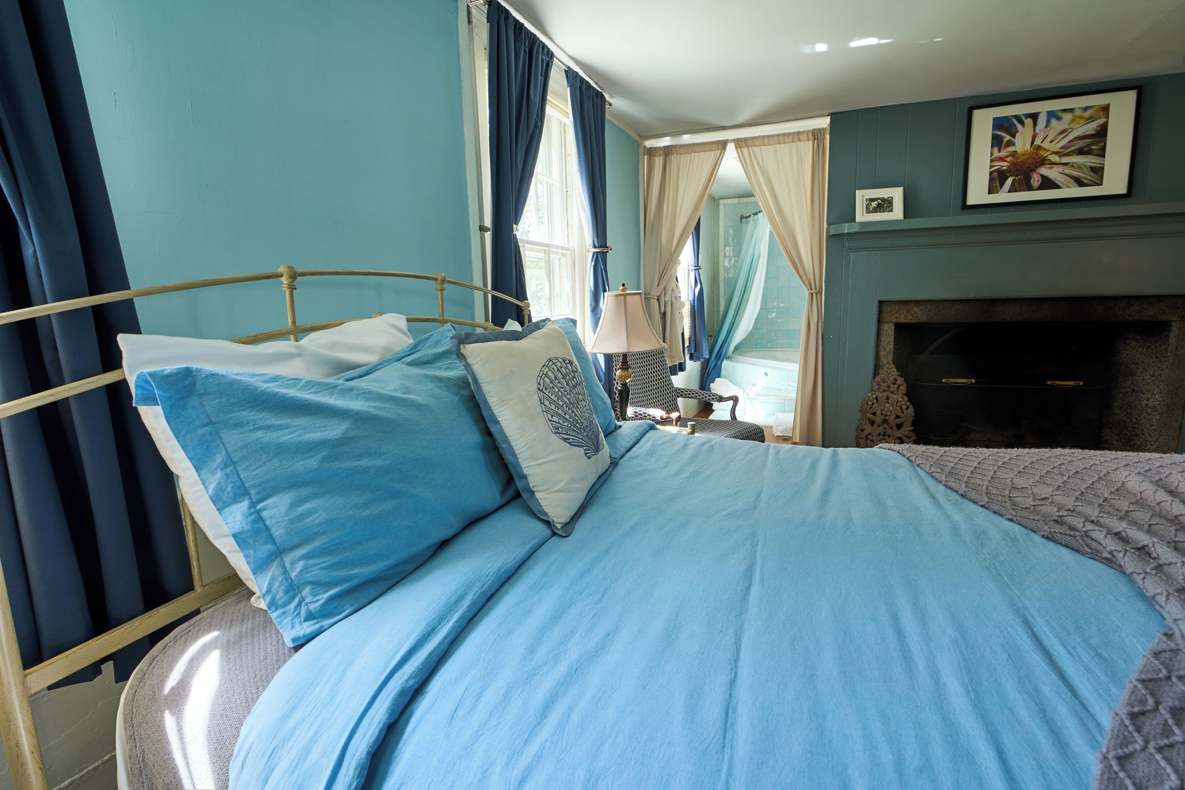A bedroom with a bed, curtains, a nightstand, a lamp, a fireplace, and a framed picture on the wall.