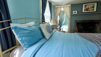 A bedroom with a bed, curtains, a nightstand, a lamp, a fireplace, and a framed picture on the wall.