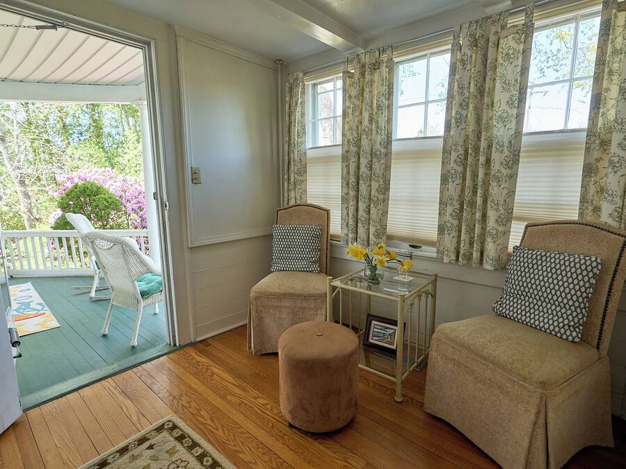 A room with a sliding glass door leading to a balcony, two upholstered chairs with patterned cushions, a small table with a vase of flowers, and a wooden floor.