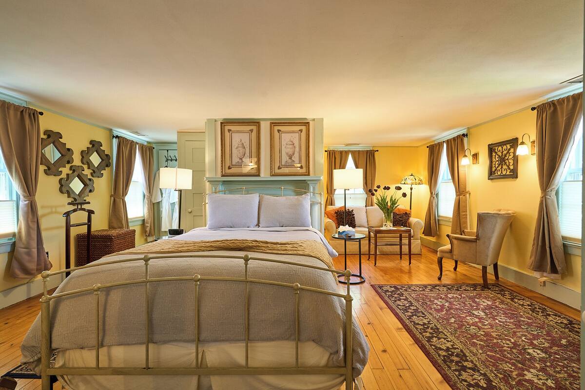 A hotel room with a large bed, a sitting area with a chair and a table, and a small side table with a lamp.