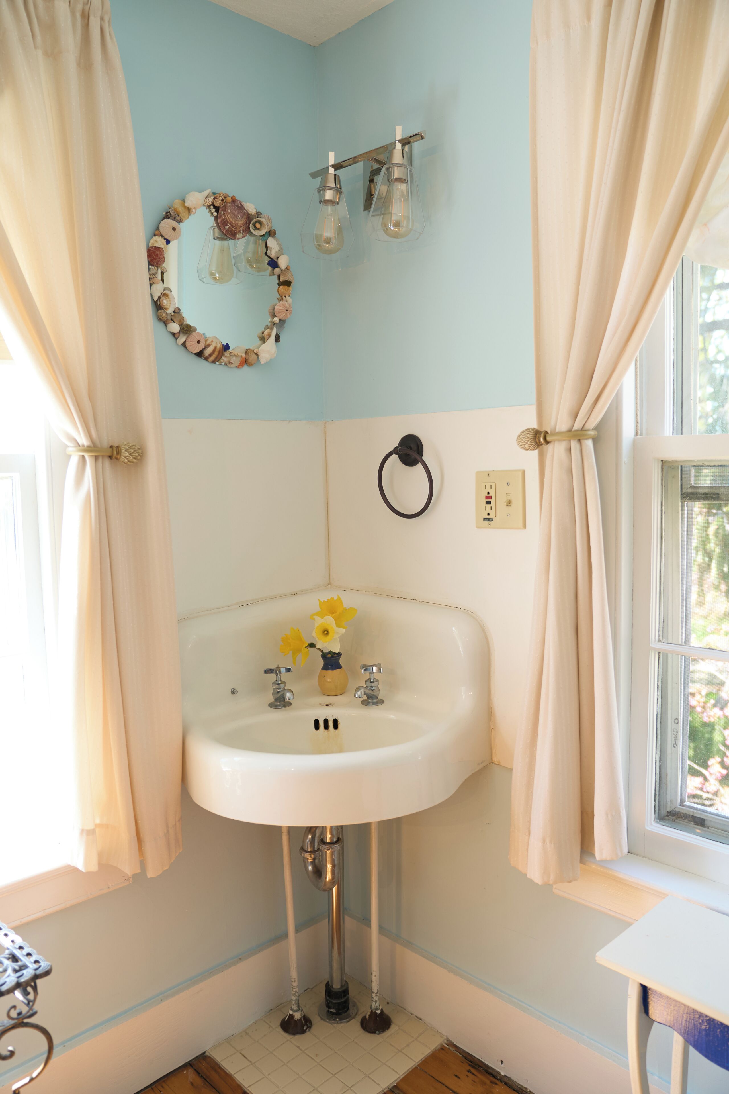 Une salle de bain avec un lavabo blanc, un miroir doté d’un cadre décoratif et une fenêtre avec des rideaux.
