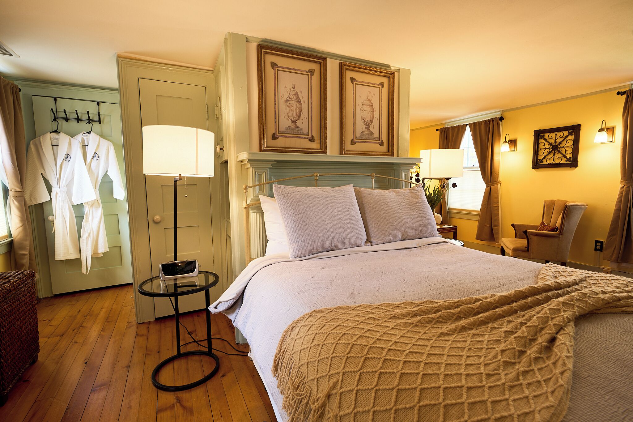 A hotel room with a bed, bedside lamps, a small table, a chair, and a wardrobe with robes.