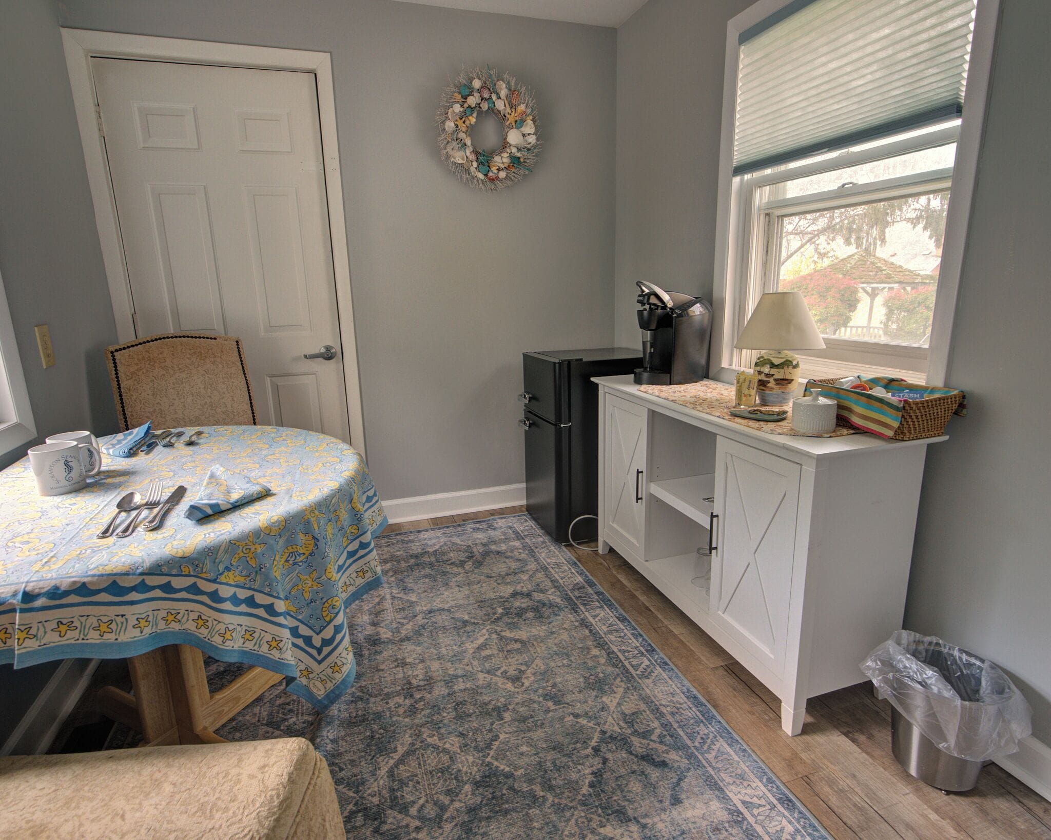 A dining area with a table set for two, a small cabinet, a waste bin, and a window with blinds.