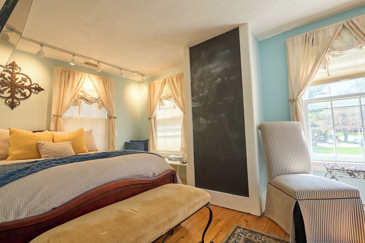 A bedroom with a bed, a chair, a large window, and a wall-mounted artwork.