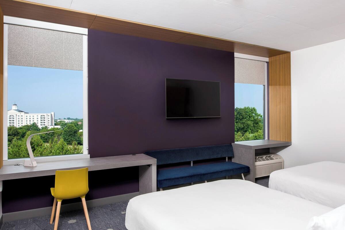 A modern hotel room with a large window, a desk, a chair, a TV, and two beds.