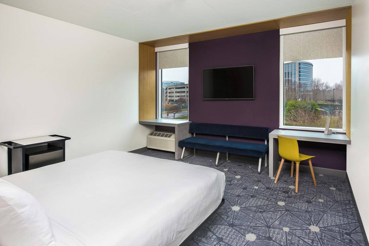 A hotel room with a bed, a desk, a chair, a TV, and a window with a view of buildings.