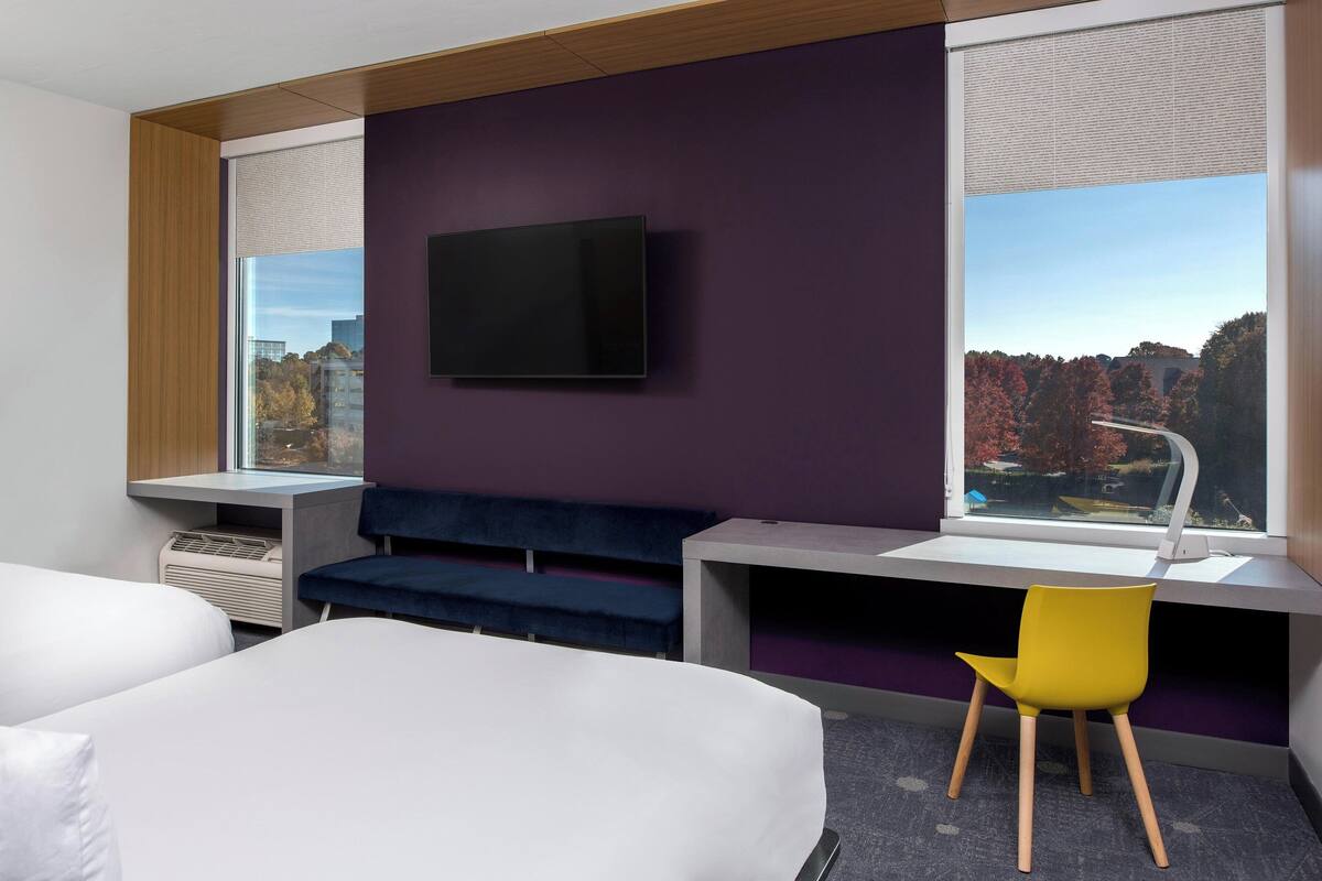 A modern hotel room with a large bed, a desk with a chair, a flat-screen TV mounted on the wall, and a window with a city view.