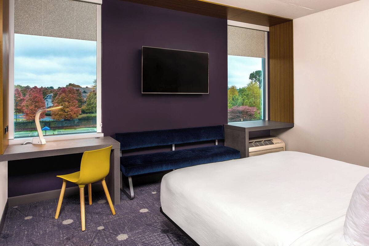 A modern hotel room with a large bed, a desk with a chair, a flat-screen TV mounted on the wall, and a view of trees and a lake through the windows.