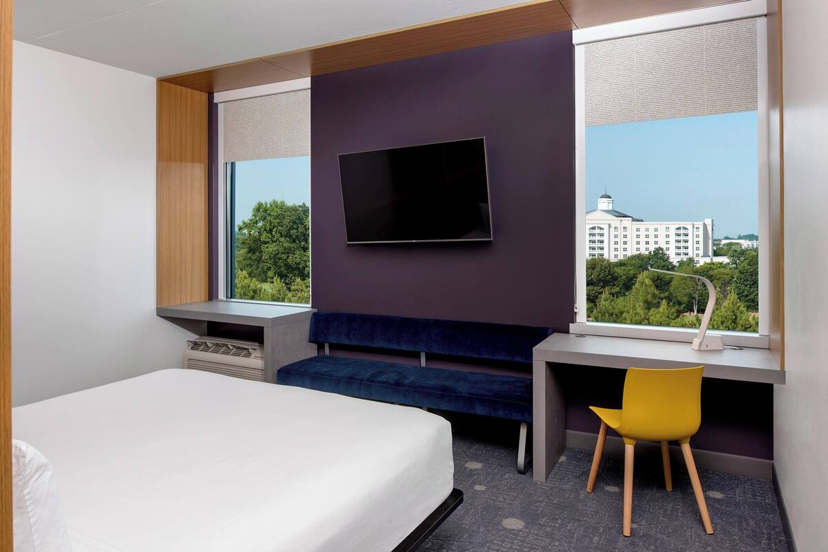 A hotel room with a bed, a desk, a chair, a TV, and a window with a city view.