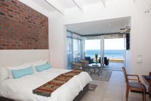 Un dormitorio con una cama grande, una pared de ladrillo, un balcón con vistas a la playa y una zona de comedor con mesa y sillas.