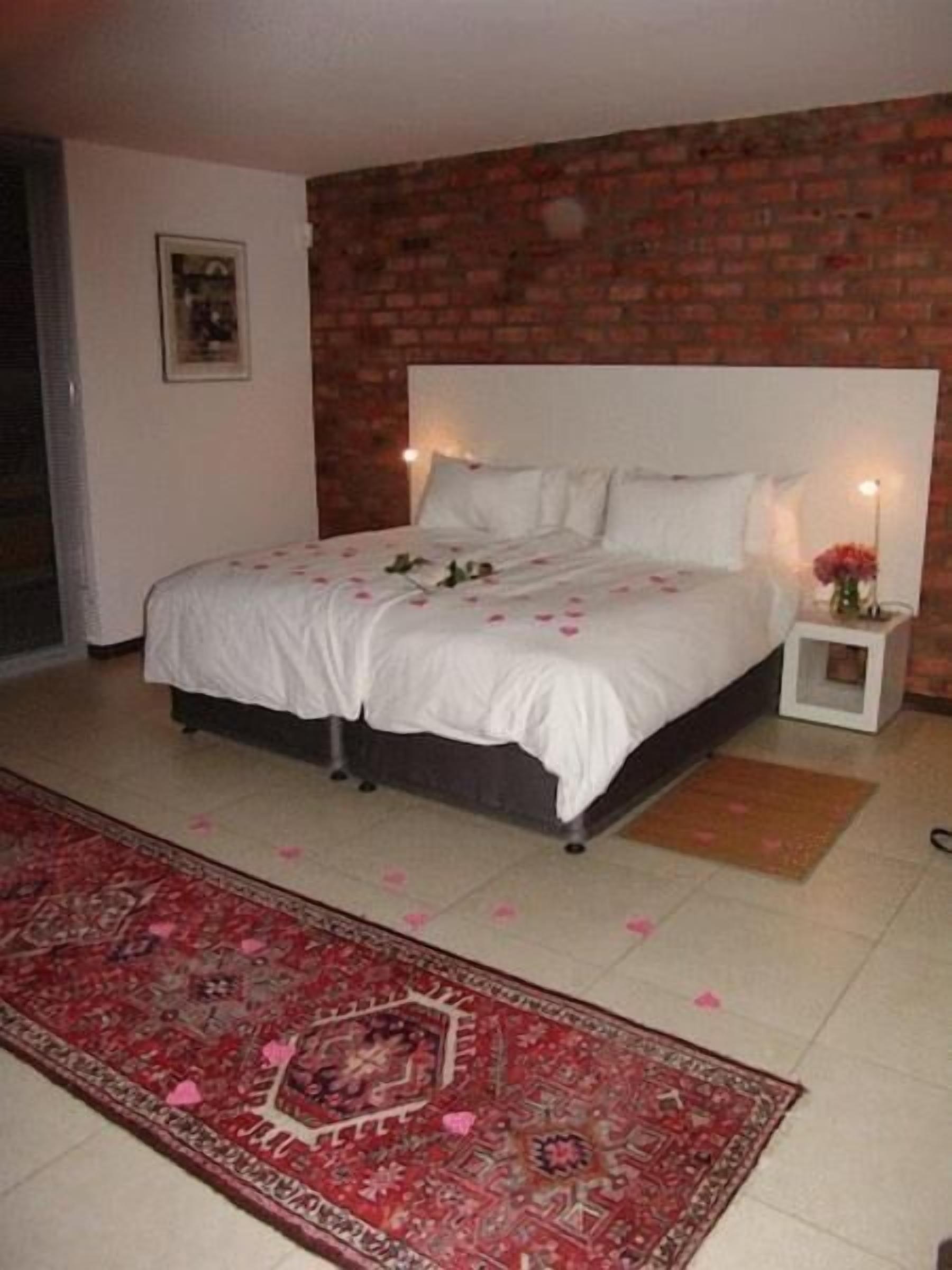 A bedroom with a brick wall, a large bed with white bedding, a nightstand with a lamp, and a red patterned rug.