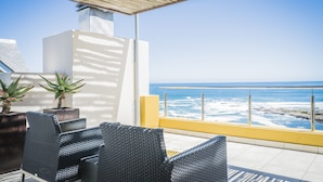 Uma varanda com cadeiras de vime, guarda-corpo amarelo e vista para o mar.