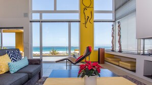 Uma sala de estar espaçosa com uma grande janela com vista para a praia, um sofá confortável e uma mesa de centro com uma planta em vaso.