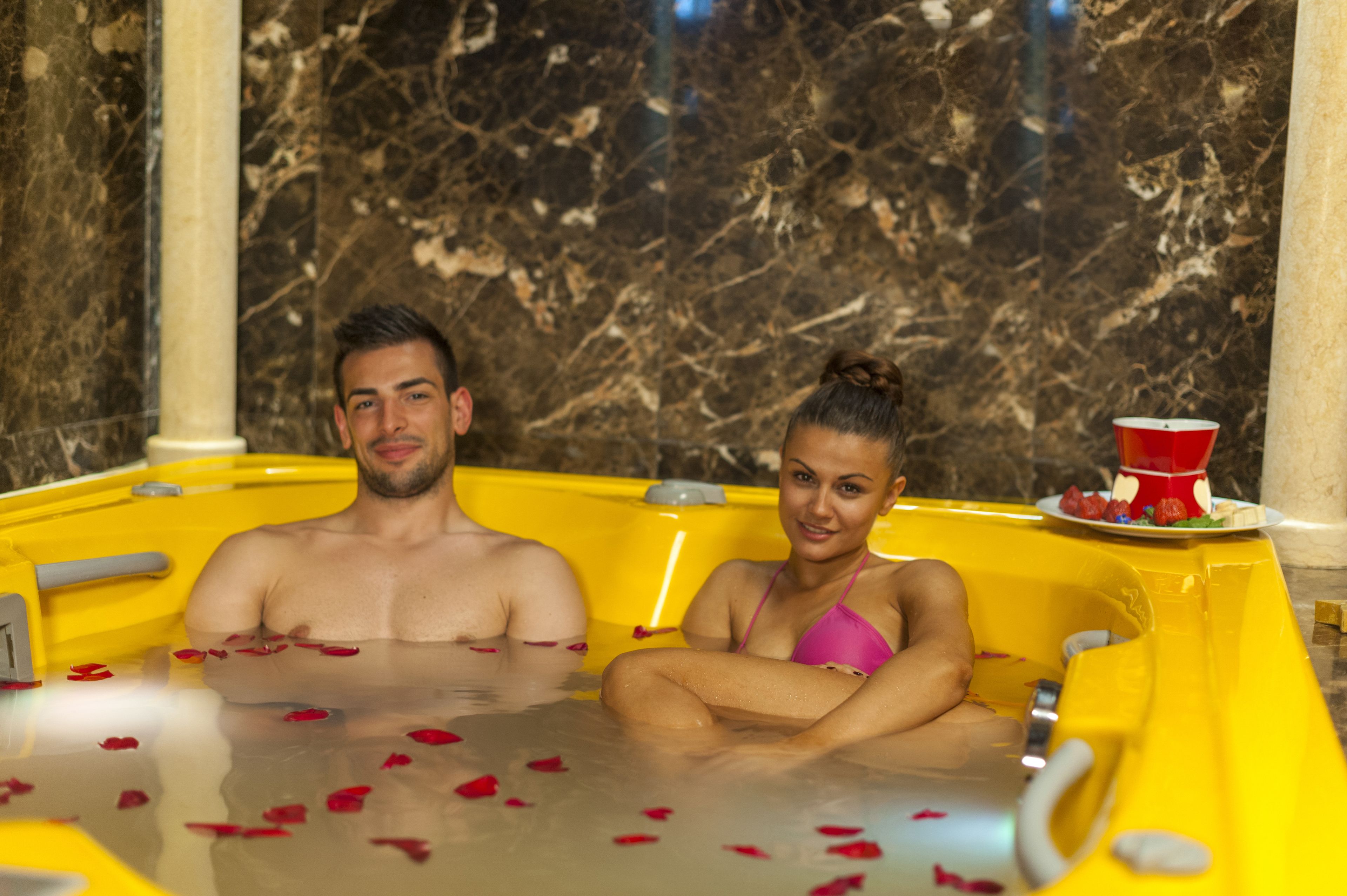 Ein Paar in einer gelben Badewanne mit Rosenblättern, Marmorwänden und einer roten Tasse auf einem Tablett.