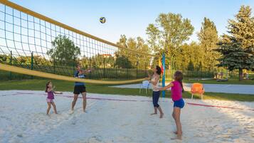 A volleyball game on a sandy court with a net, surrounded by trees and a fence.