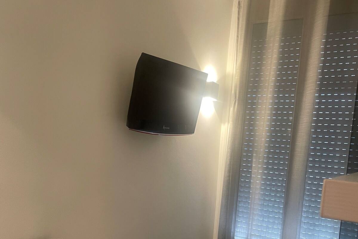 A hotel room with a bed, a desk with a lamp, a flat-screen TV mounted on the wall, and a window with blinds.