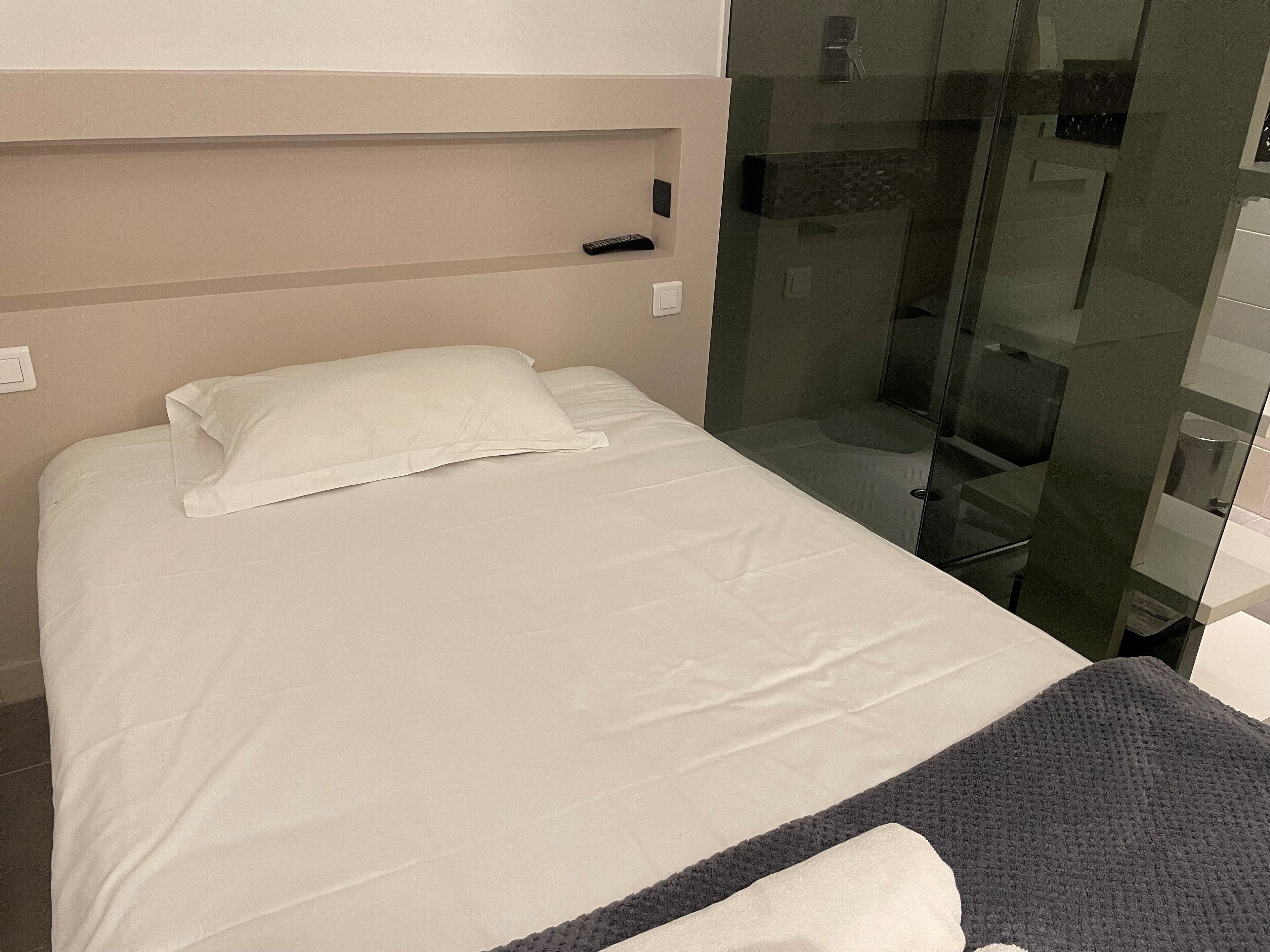 A neatly made bed with white linens and a gray blanket, a glass shower enclosure, and a white wall with a light switch.