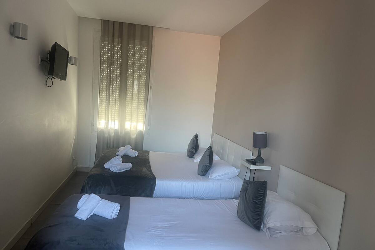A hotel room with two beds, a TV mounted on the wall, a window with curtains, and a bedside table with a lamp.