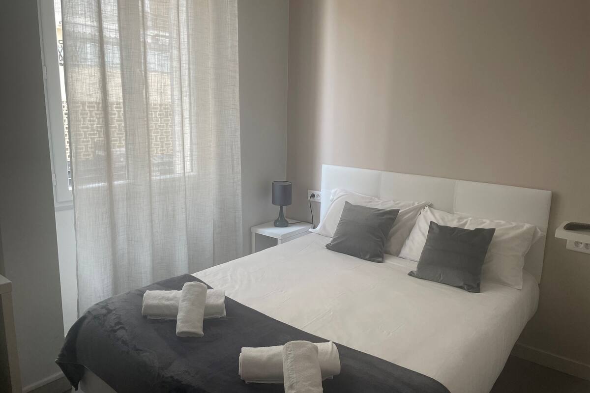 A neatly made bed with white and gray linens, a bedside lamp, and a window with sheer curtains.