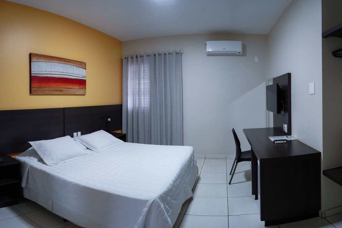 Een hotelkamer met een bed, een bureau, een stoel, een airconditioning, een televisie en een schilderij aan de muur.