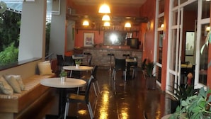 A cozy restaurant with a long counter, multiple tables, and chairs. There are hanging lights and a large window on the right side.