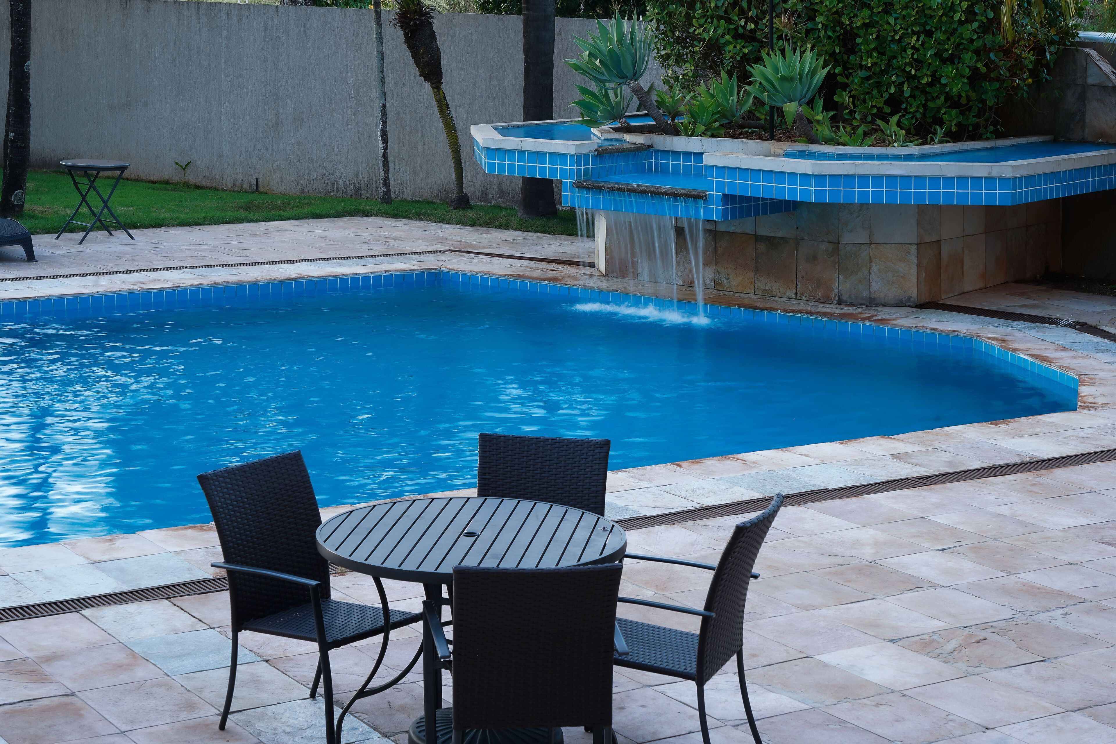 A swimming pool with a waterfall feature, surrounded by a tiled patio with a table and chairs.