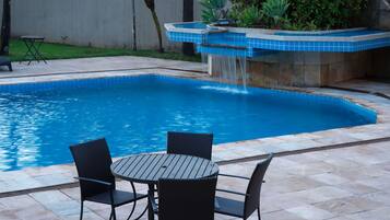 A swimming pool with a waterfall feature, surrounded by a tiled patio with a table and chairs.
