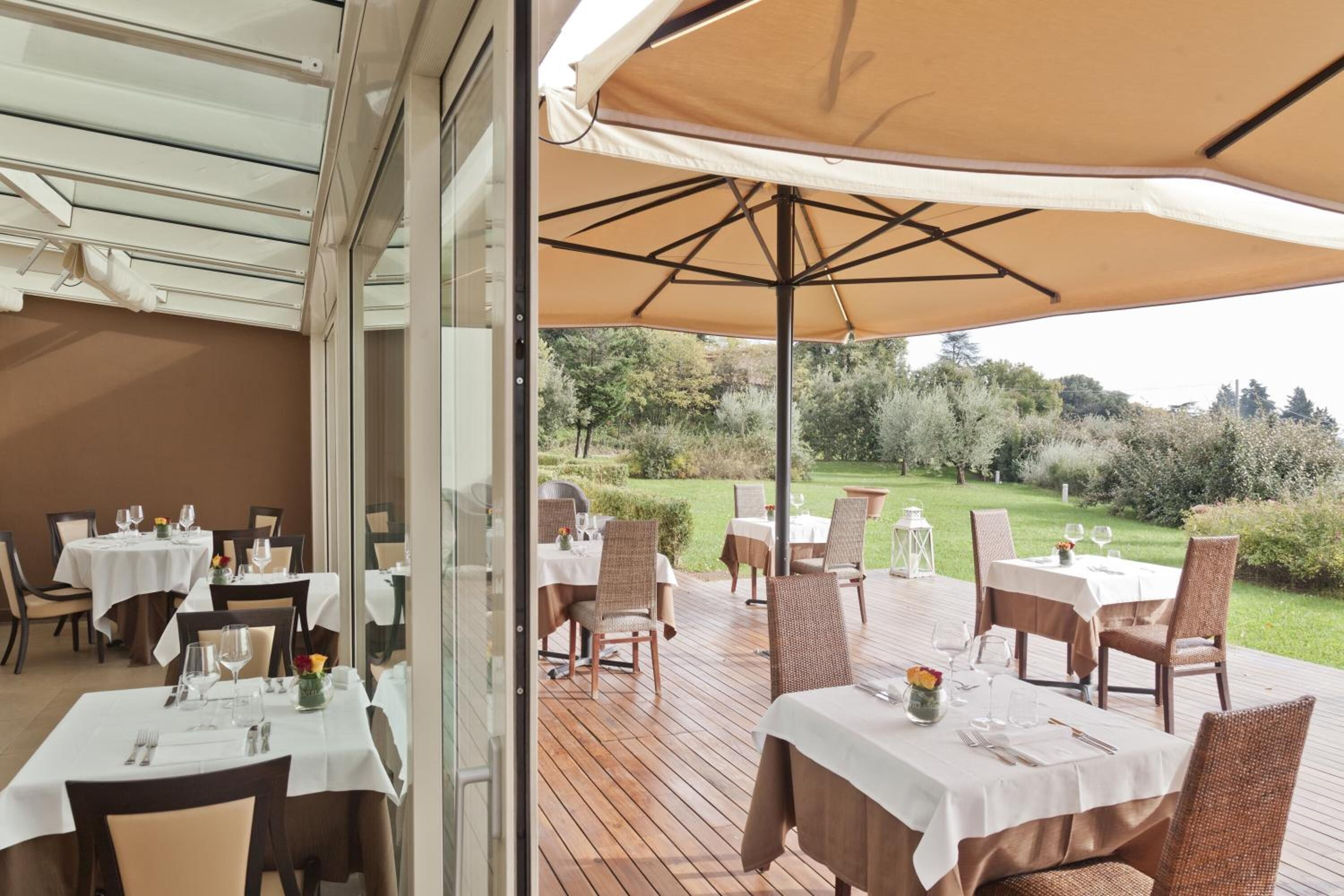 An outdoor dining area with tables and chairs, a large umbrella, and a view of a garden.