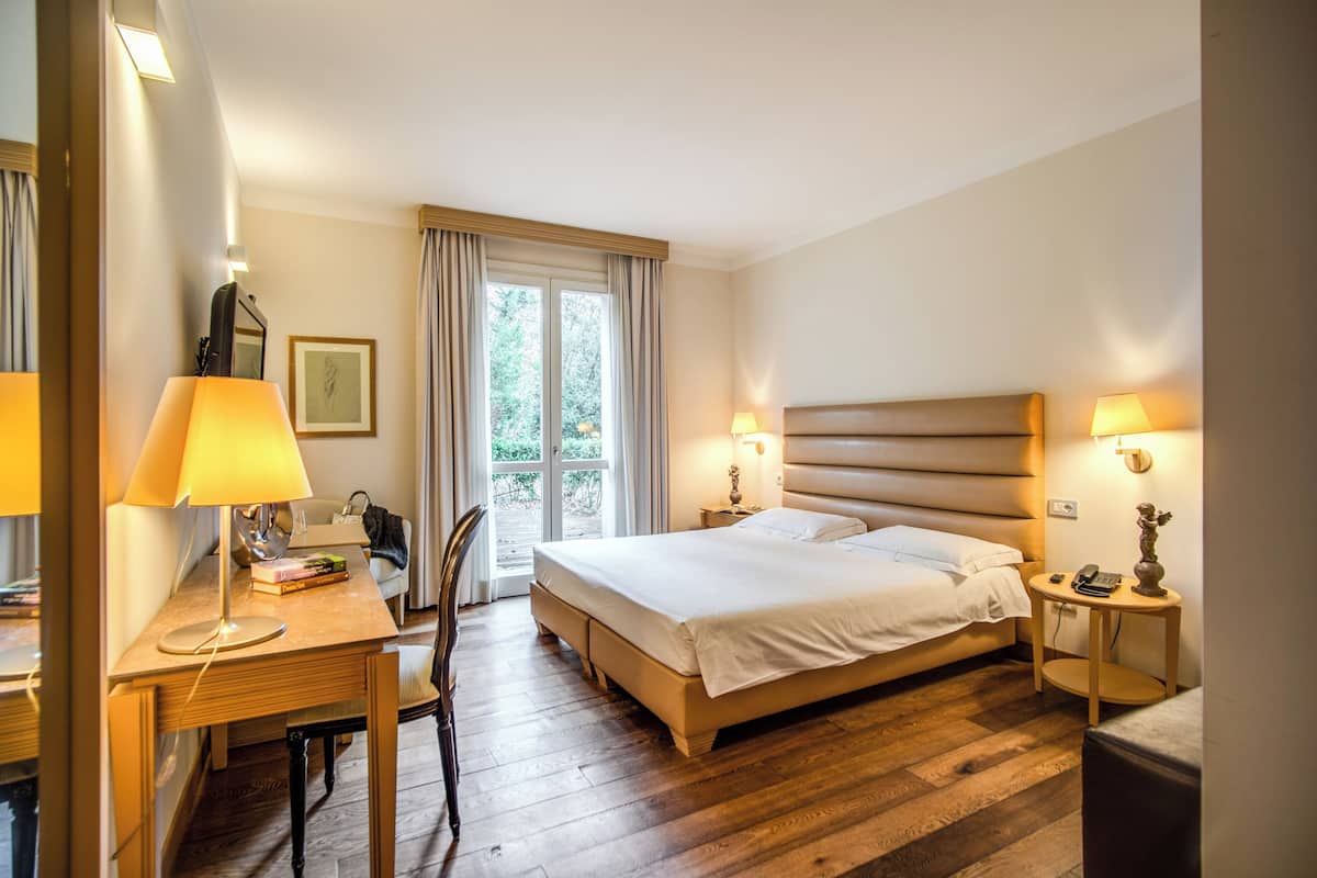 A hotel room with a bed, bedside table, desk, chair, and a view of the outdoors.