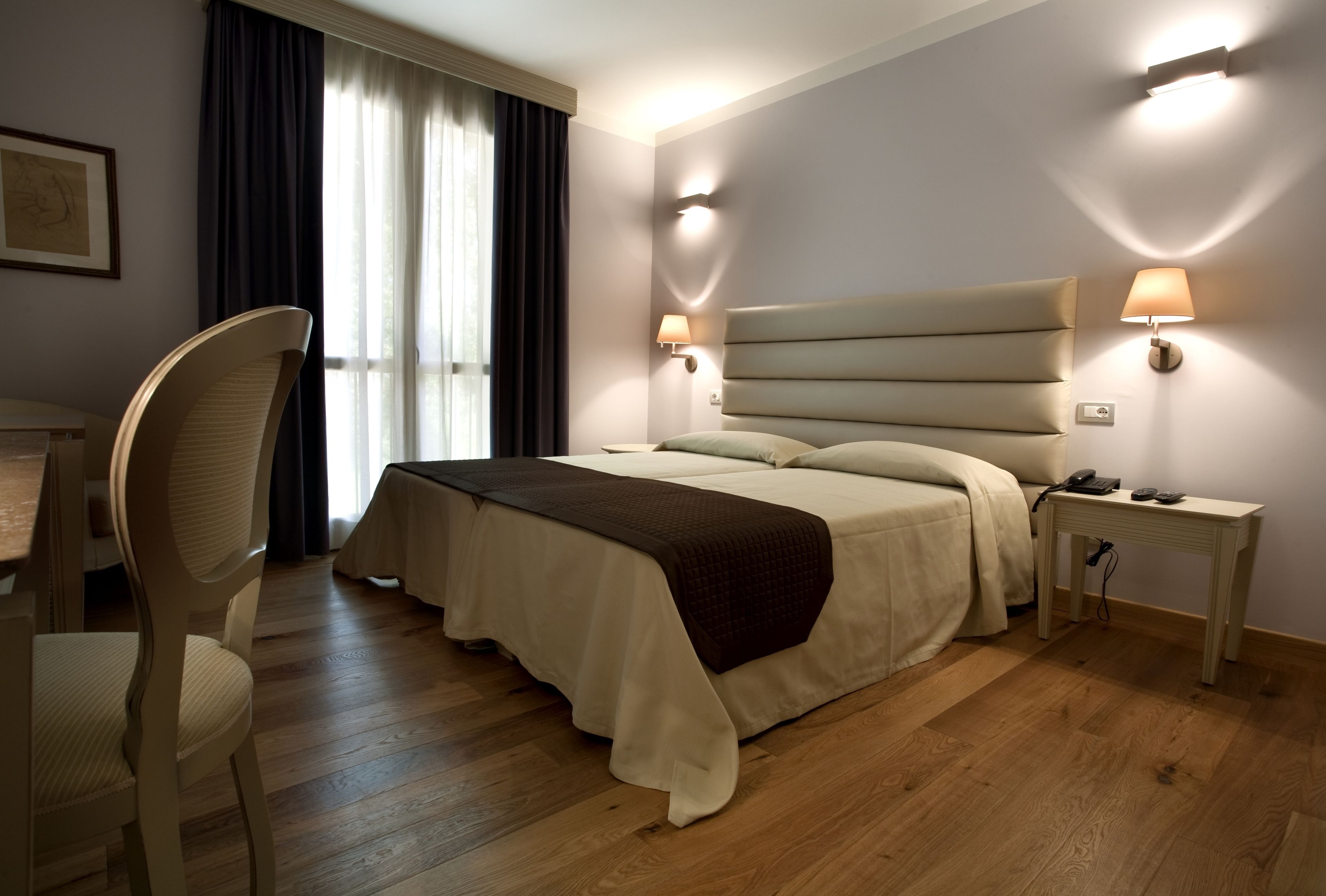Hotelkamer met een groot bed, een bureau en een stoel. Twee bedlampjes en een raam met gordijnen.