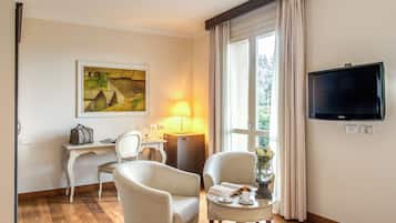 A hotel room with a wooden floor, a desk with a lamp, two chairs, a television, and a painting on the wall.