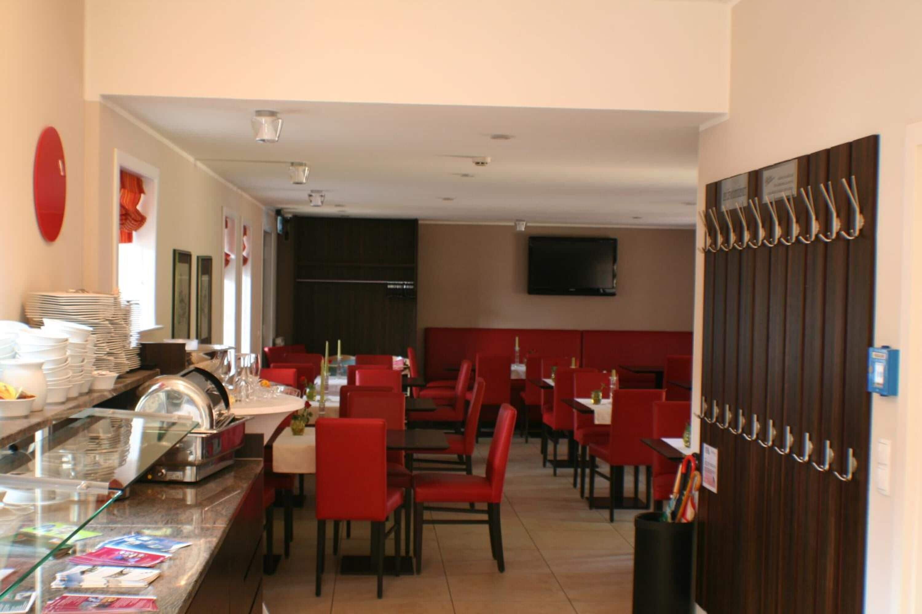 Un restaurante con sillas y mesas rojas, una zona de bufé con platos apilados y un televisor montado en la pared.