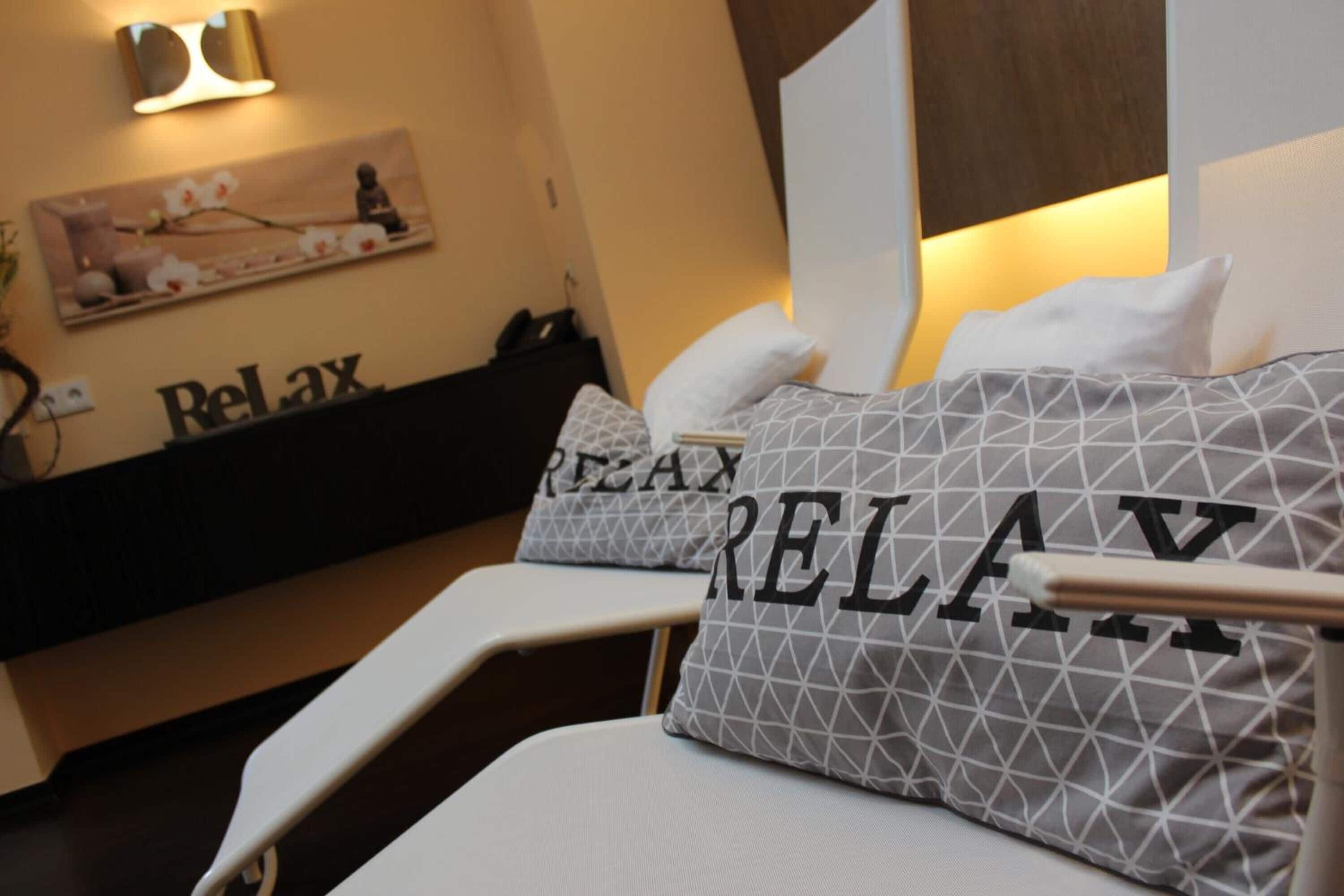 Ein Bett mit Kissen, die das Wort 'RELAX' zeigen, ein Nachttisch und eine Wand mit einem 'RELAX'-Schild und einem Blumenbild.