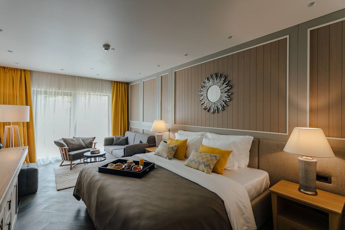 A modern hotel room with a large bed, a sitting area with a sofa and armchair, a dining table with a tray of breakfast, and a decorative mirror on the wall.