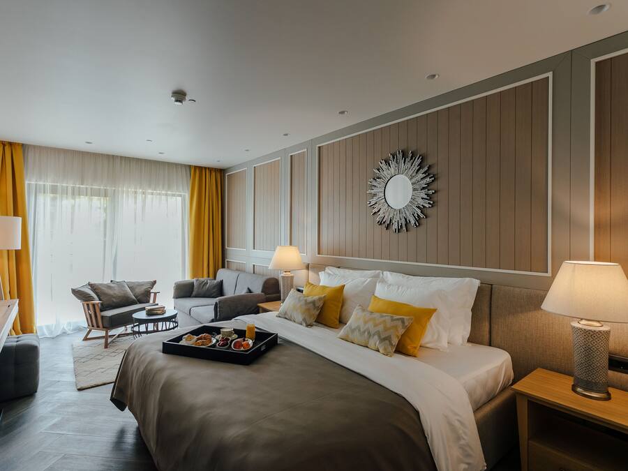 A modern hotel room with a large bed, a sitting area with a sofa and armchair, a dining table with a tray of breakfast, and a decorative mirror on the wall.