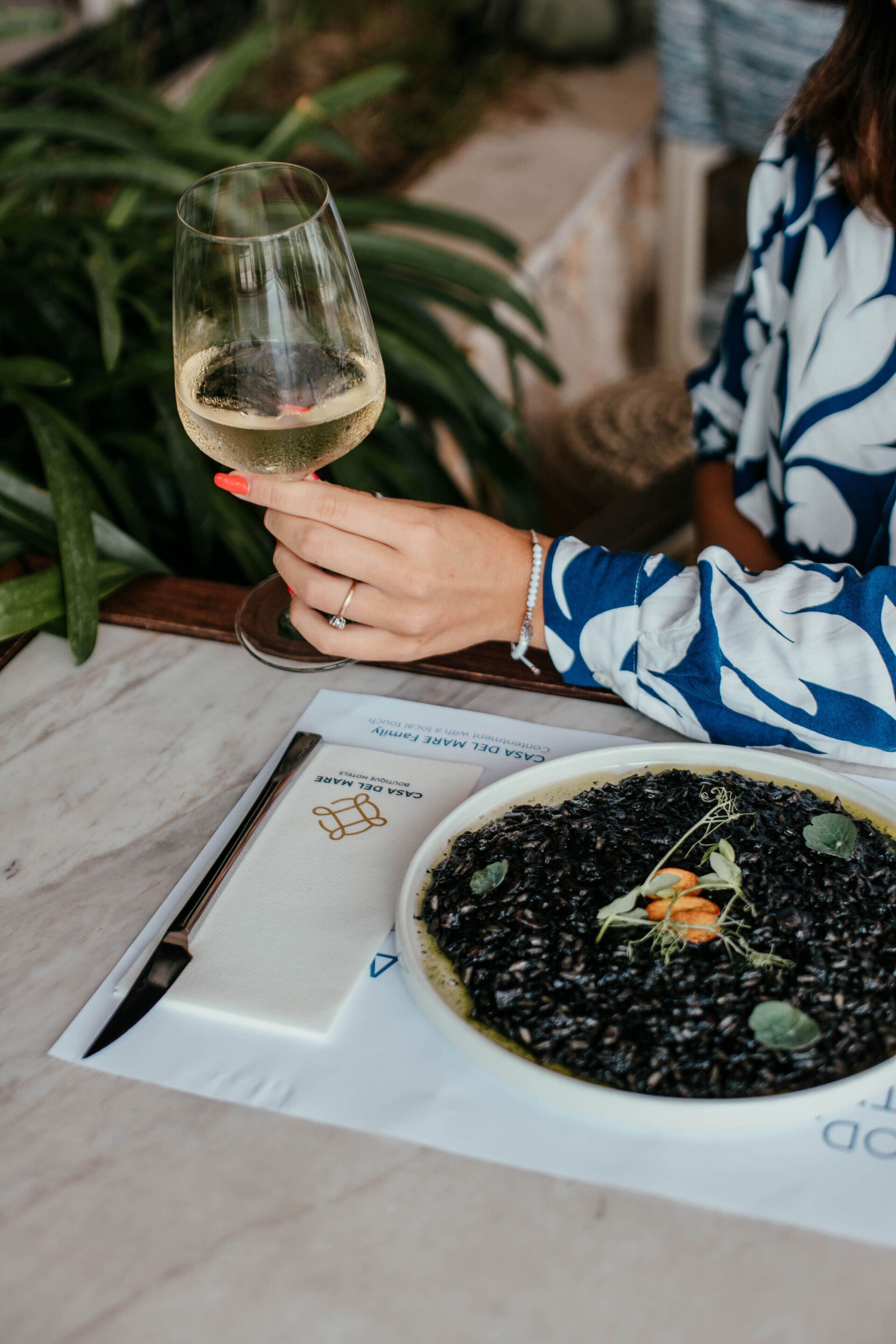 A person holding a wine glass, a menu, and a bowl of black rice with greens.