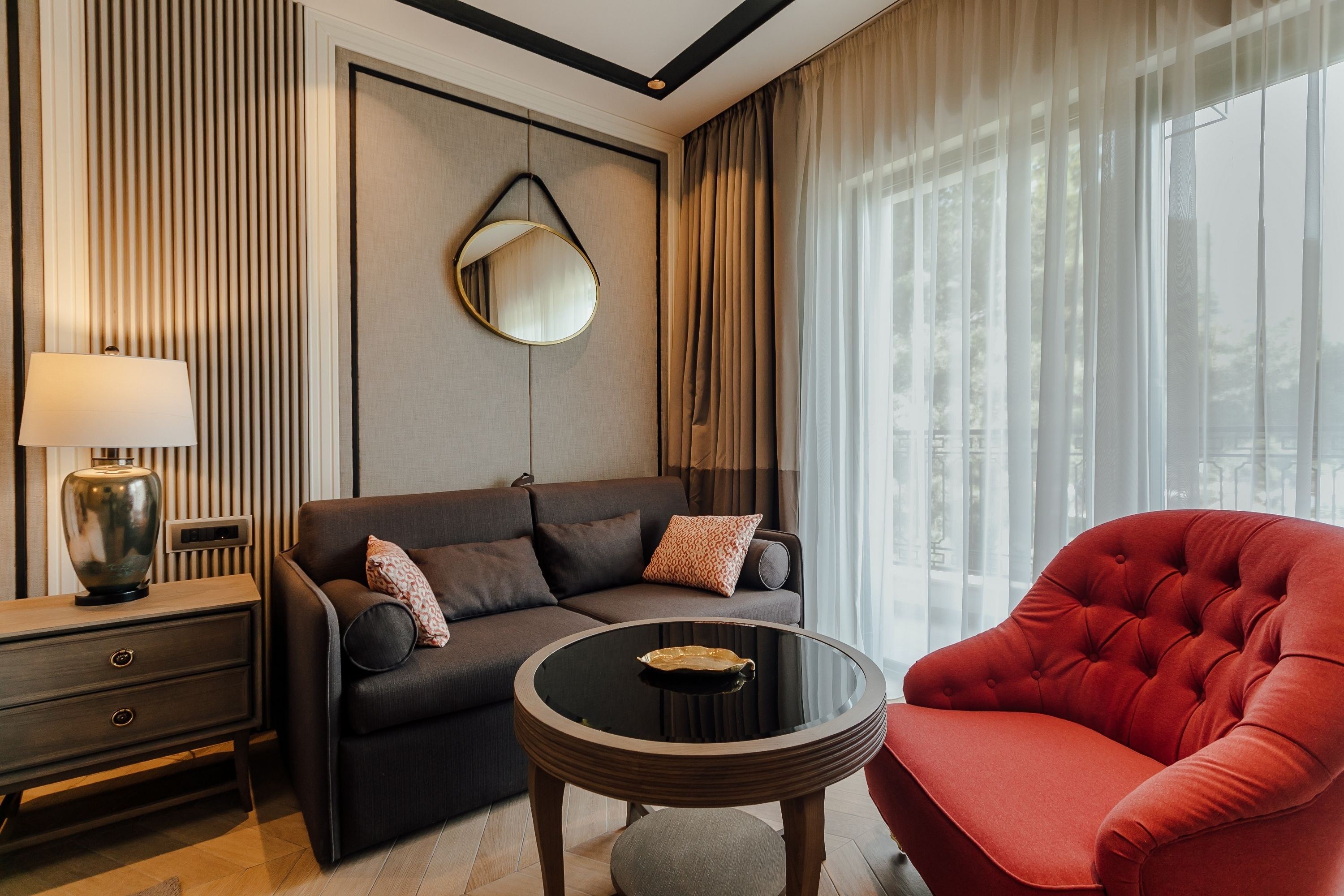 Una sala de estar moderna con un sofá, un sillón rojo, una mesa redonda, una lámpara y un espejo adosado a la pared.