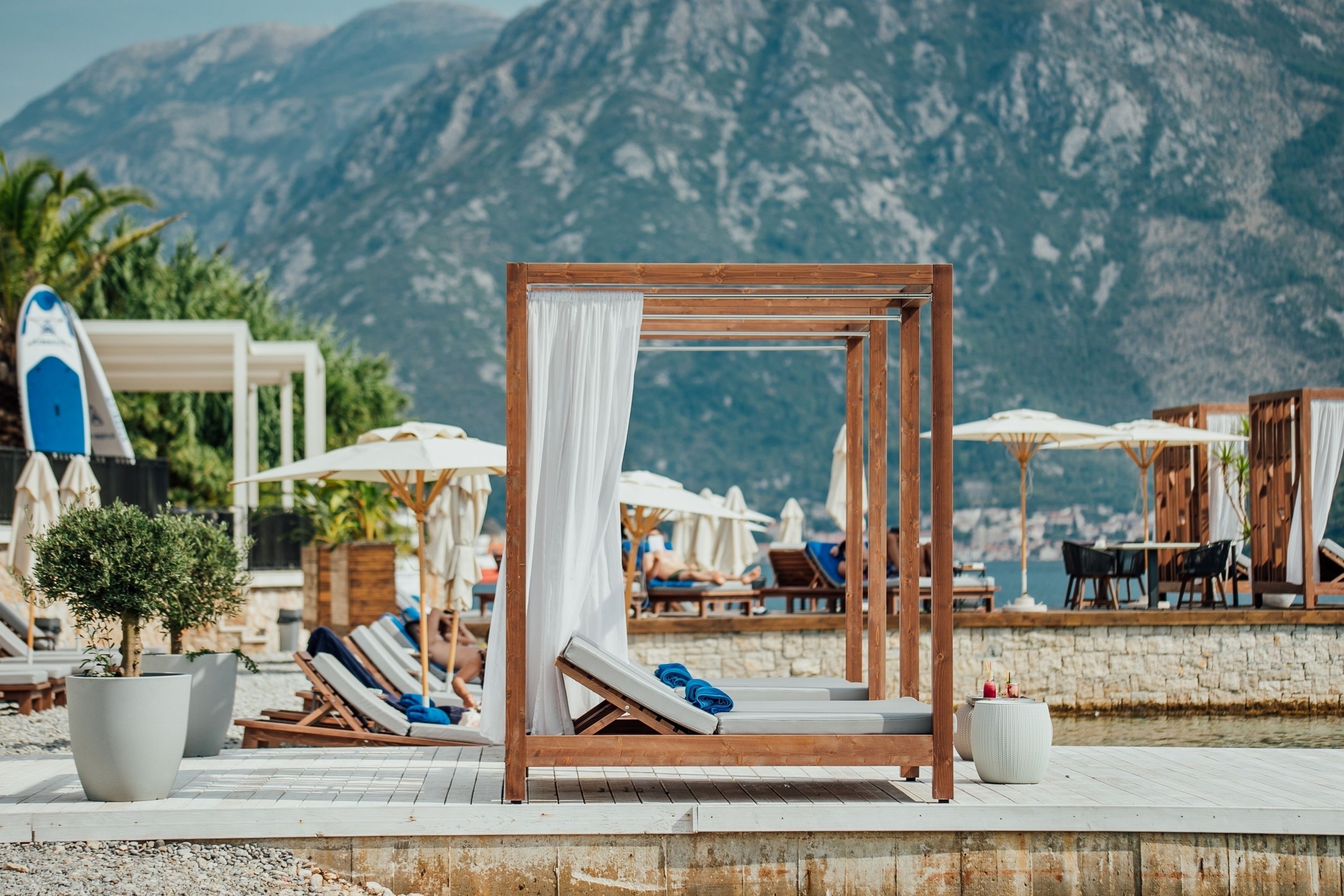 A beachside lounge area with wooden loungers, white canopies, and a view of mountains.