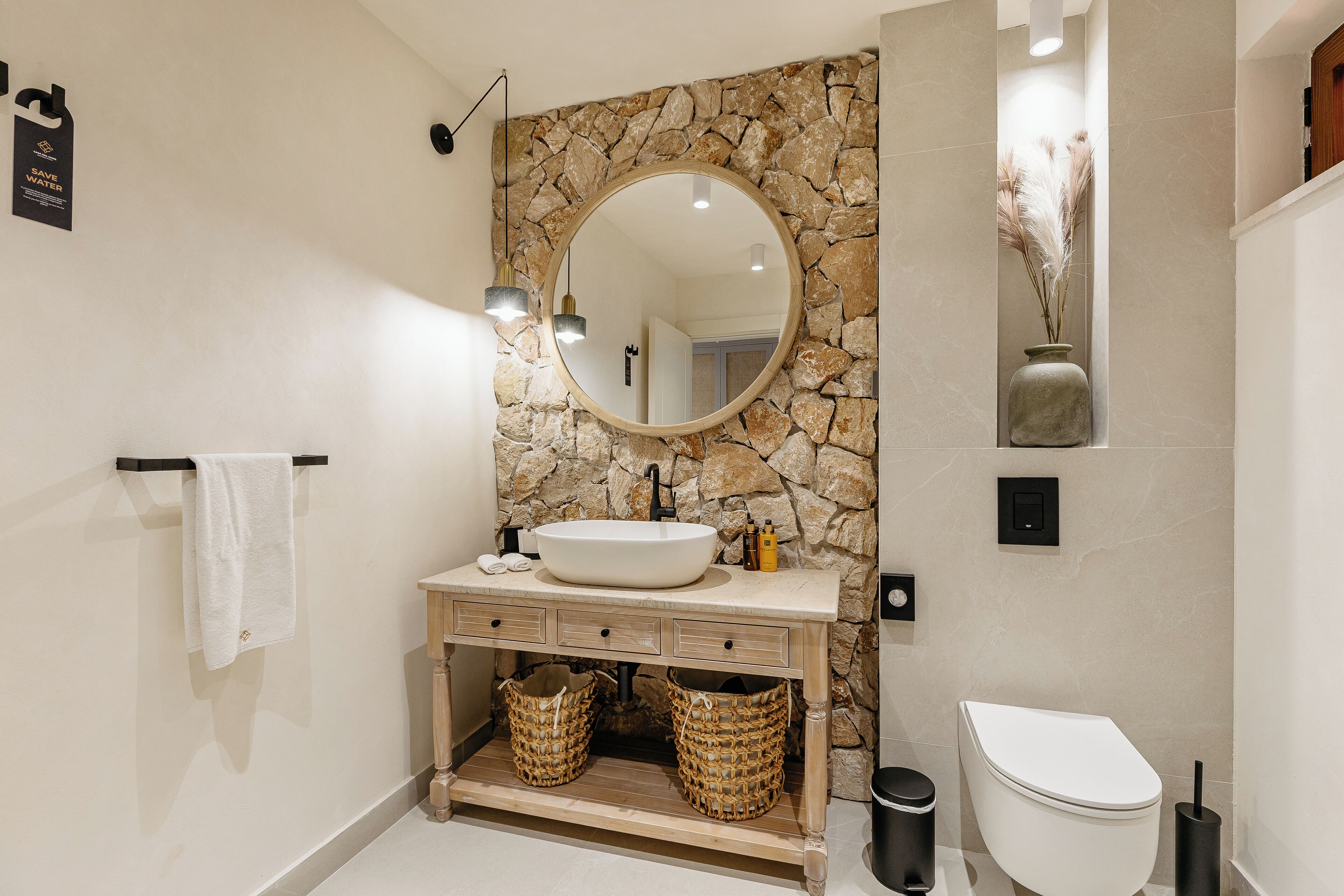 Een badkamer met een stenen muur, een ronde spiegel, een witte wastafel en een toilet.
