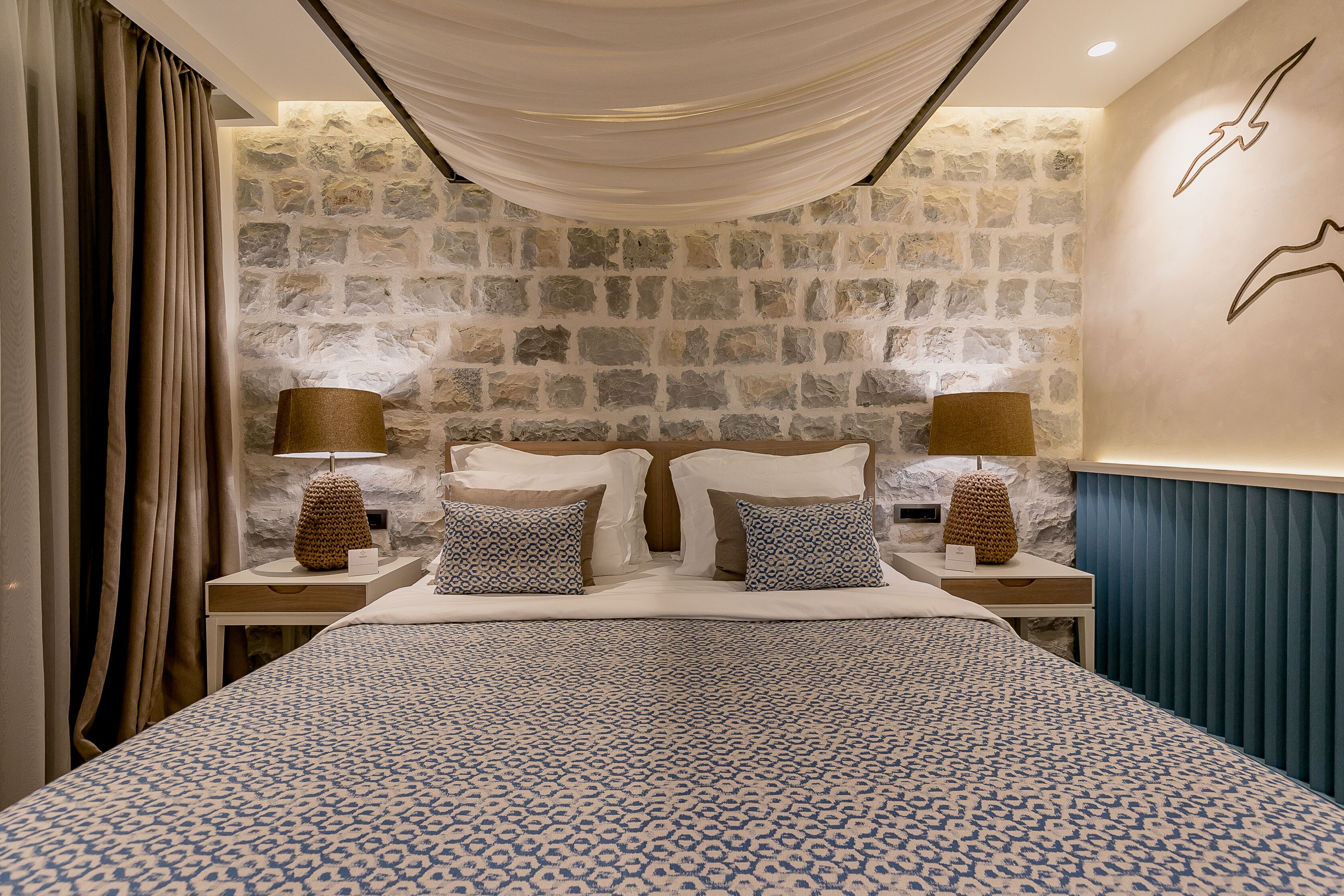 A bedroom with a stone wall, a bed with patterned bedding, two bedside tables with lamps, and a curtain on the left.