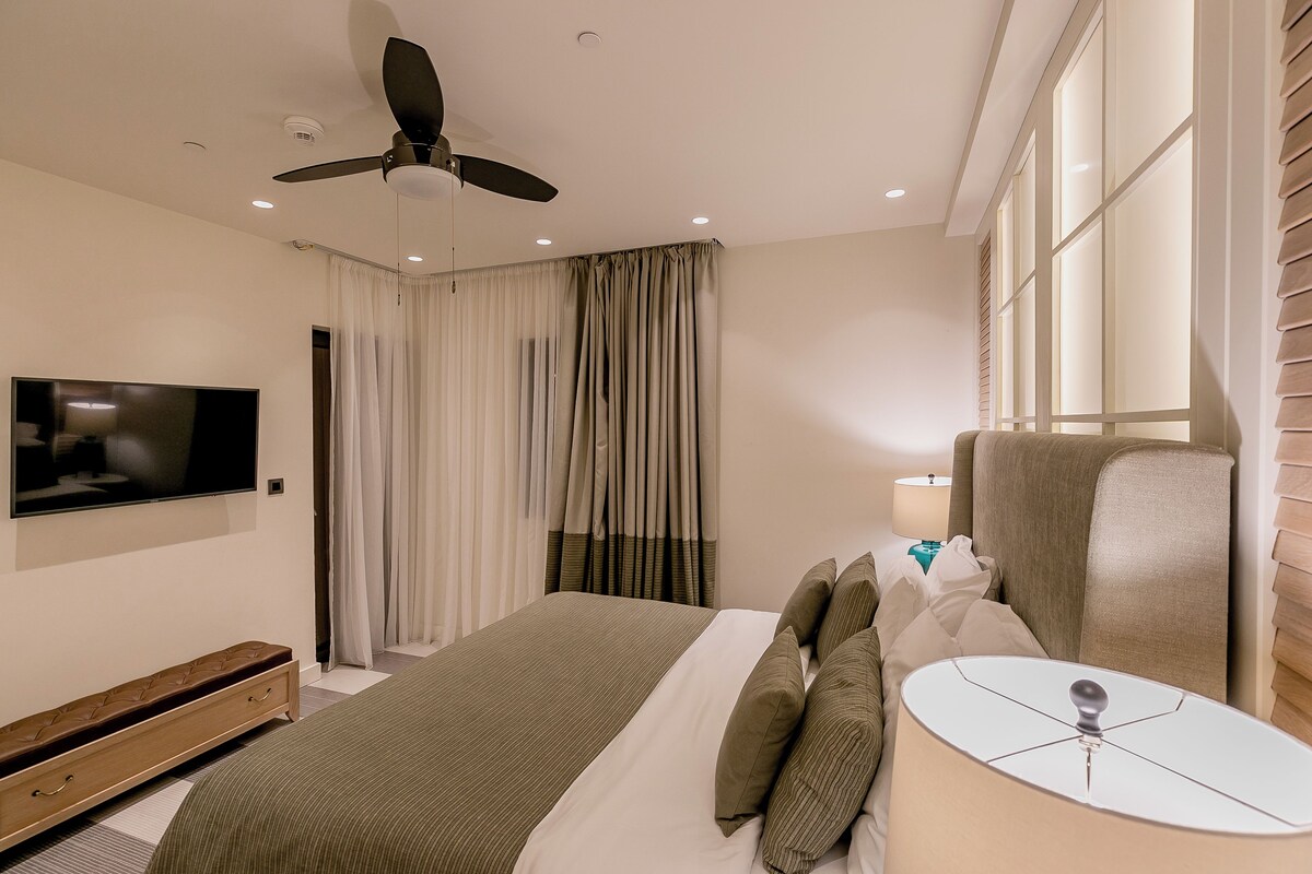 Habitación de hotel con cama, televisor, ventilador de techo, ventana con persianas y mesita de noche con lámpara.