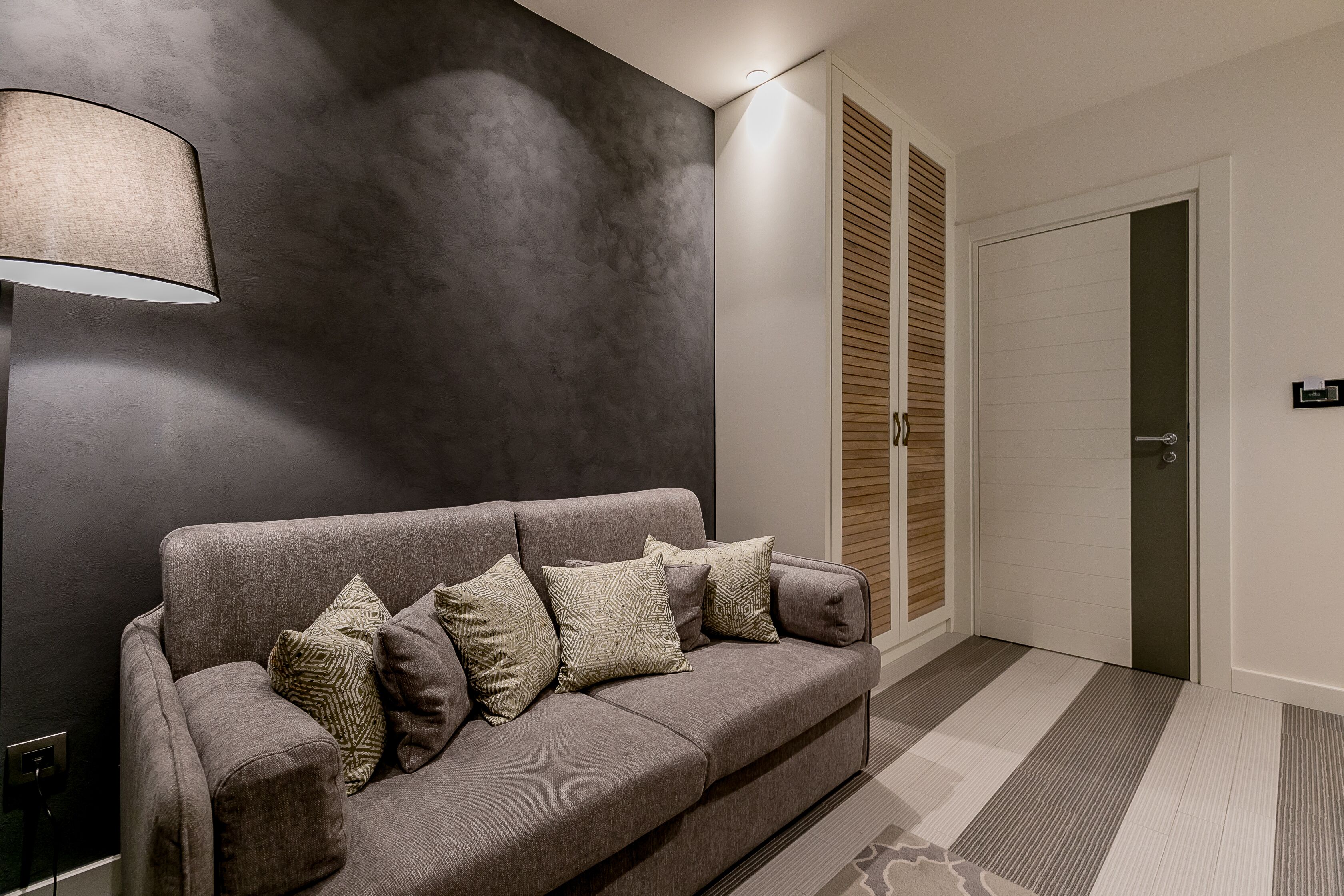 Een moderne woonkamer met een grijze bank, decoratieve kussens, een vloerlamp en een schuifdeurkast.