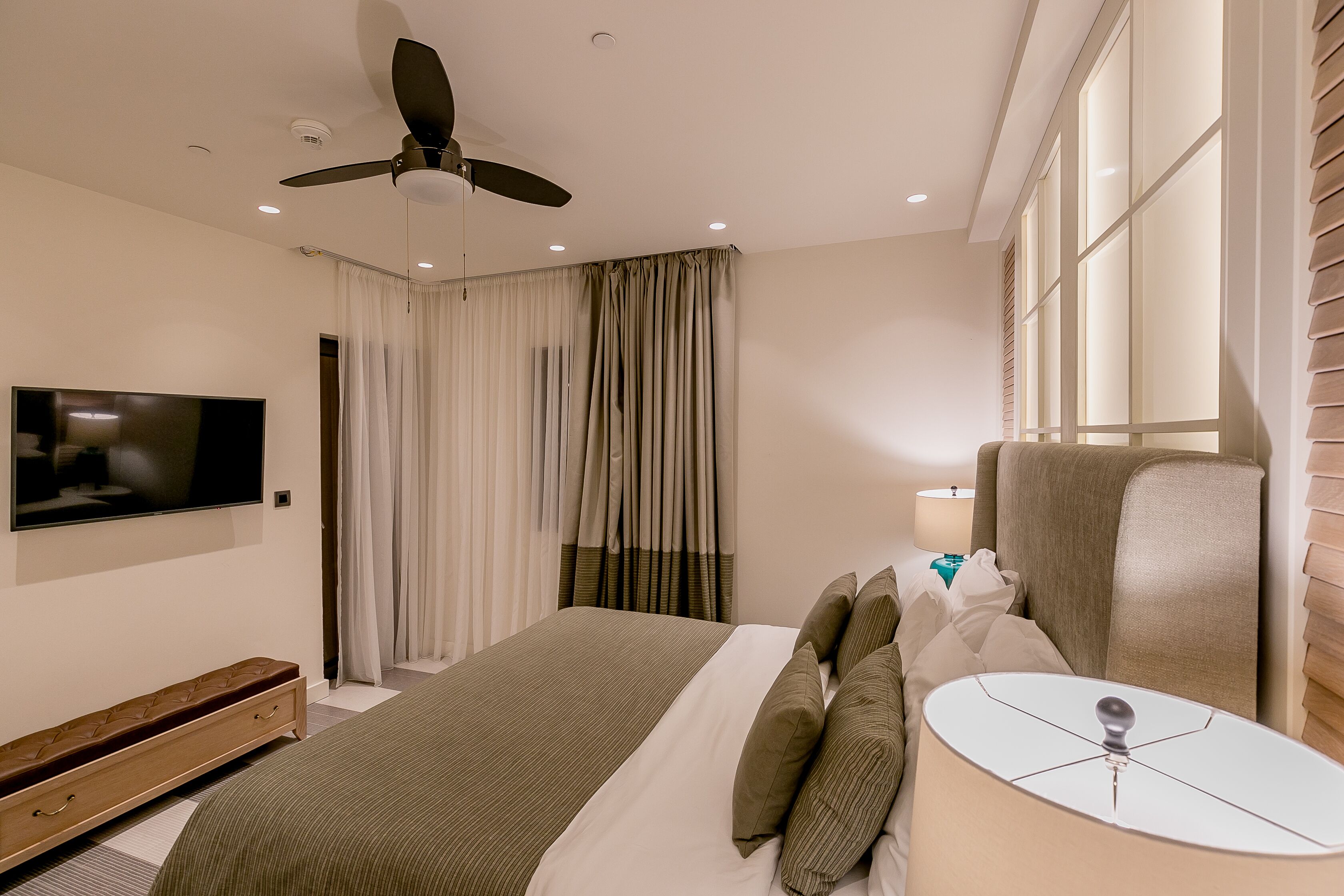 A hotel room with a bed, a television, a ceiling fan, a window with blinds, and a bedside table with a lamp.