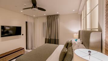 A hotel room with a bed, a television, a ceiling fan, a window with blinds, and a bedside table with a lamp.