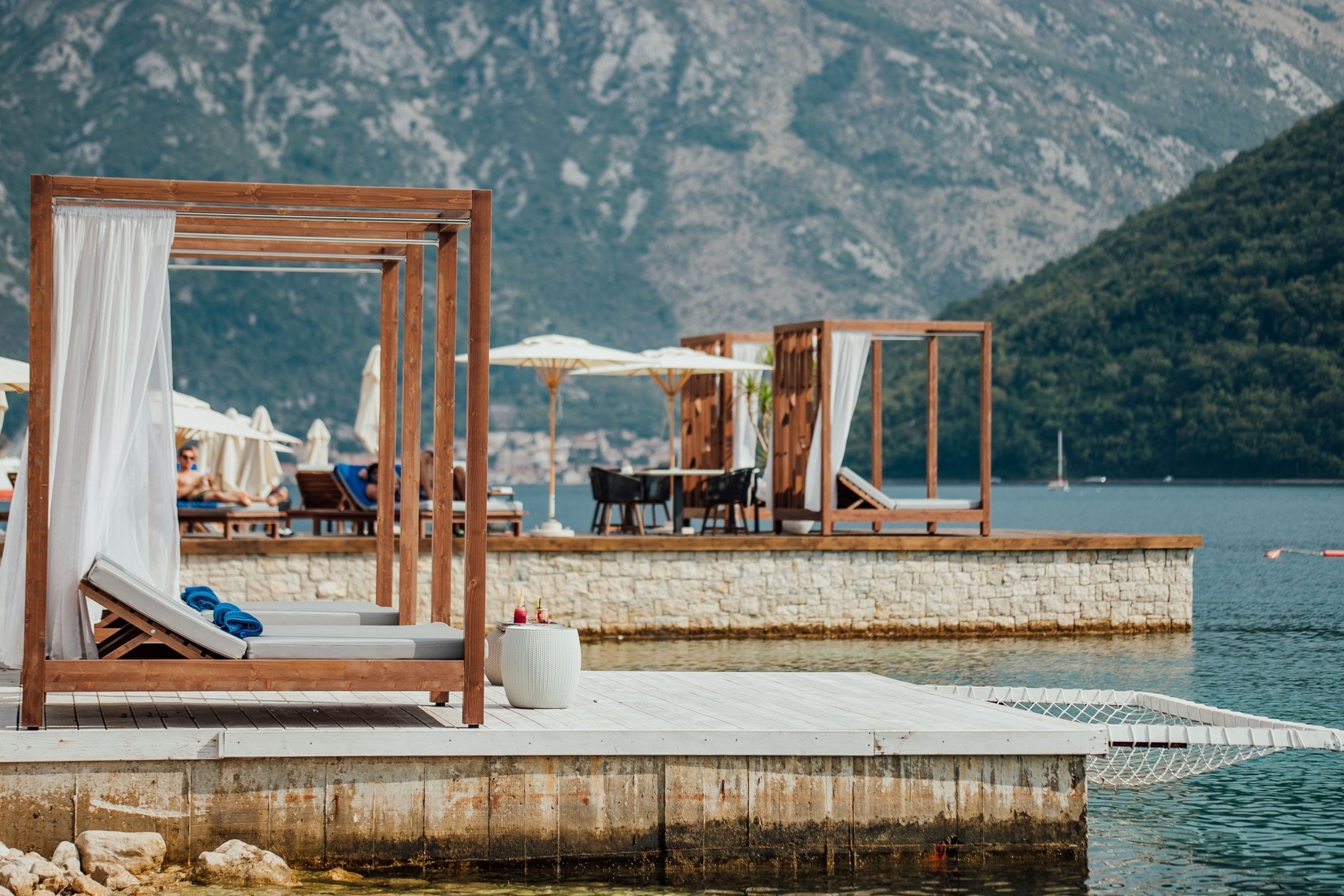 A wooden deck with lounge chairs and umbrellas, a stone wall, and a view of a lake and mountains.