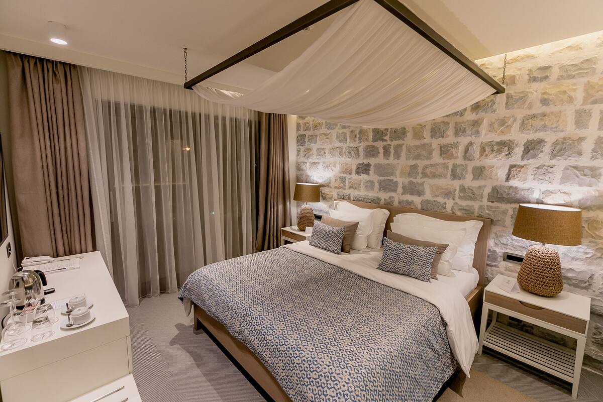 A bedroom with a large bed, a nightstand with a lamp, a dresser, and a stone wall.