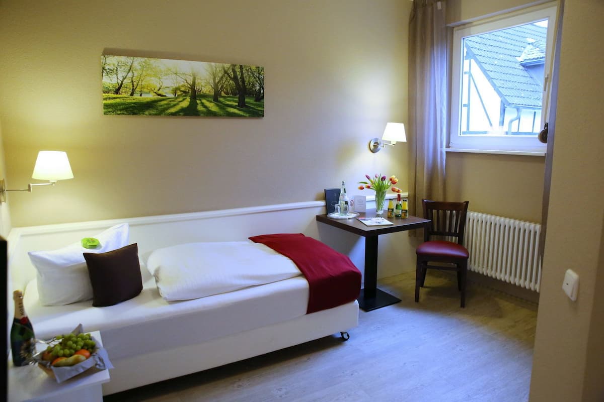 A hotel room with a bed, a small table with a vase of flowers, a chair, a radiator, and a window with a view of a building.