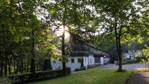 A white building with a black roof, surrounded by lush green trees and a well-maintained lawn. - Waldhotel Wilhelmshöhe (Freudenberg)
